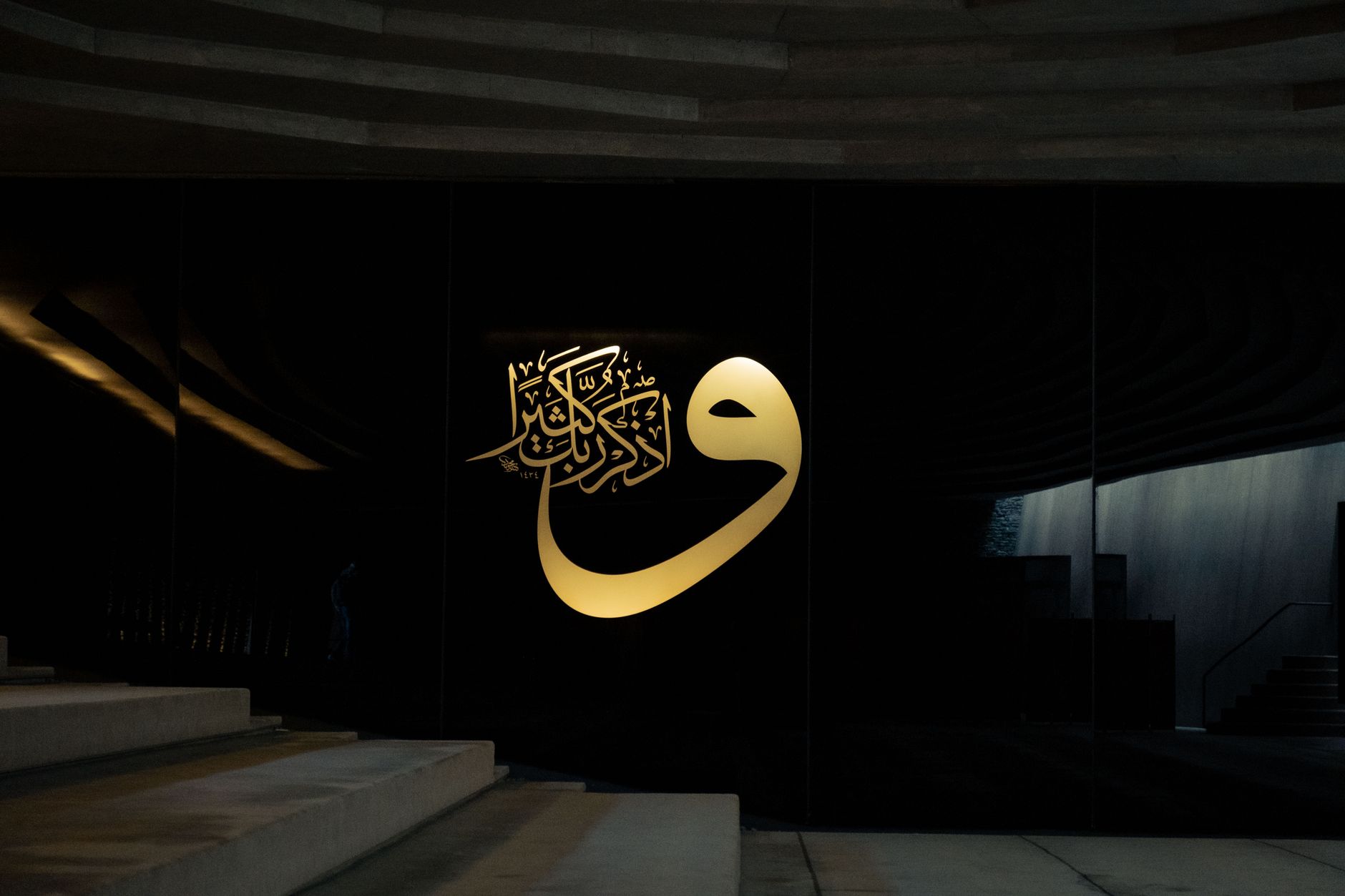 Arabic Calligraphy — Elegant Islamic calligraphy in gold on a modern black wall, featuring architectural elements and stairs.