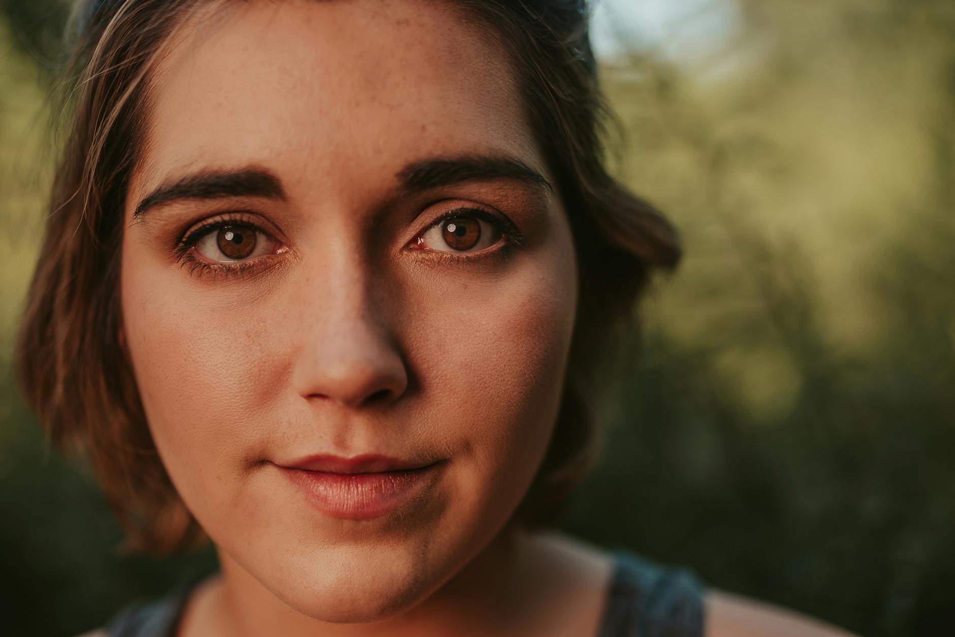 Headshot — Closeup of positive female with short hair looking at camera while standing outside with green plants on blurred background in su