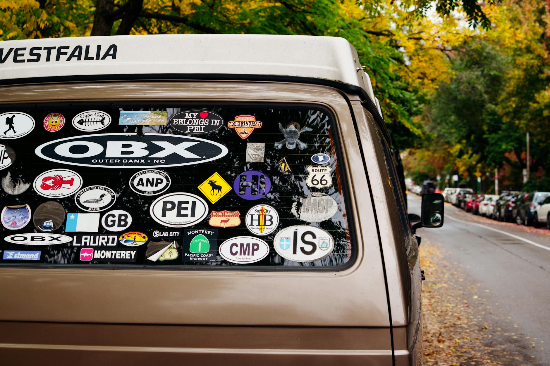 Sticker — A vintage van adorned with various travel stickers parked on an autumn street with colorful foliage.